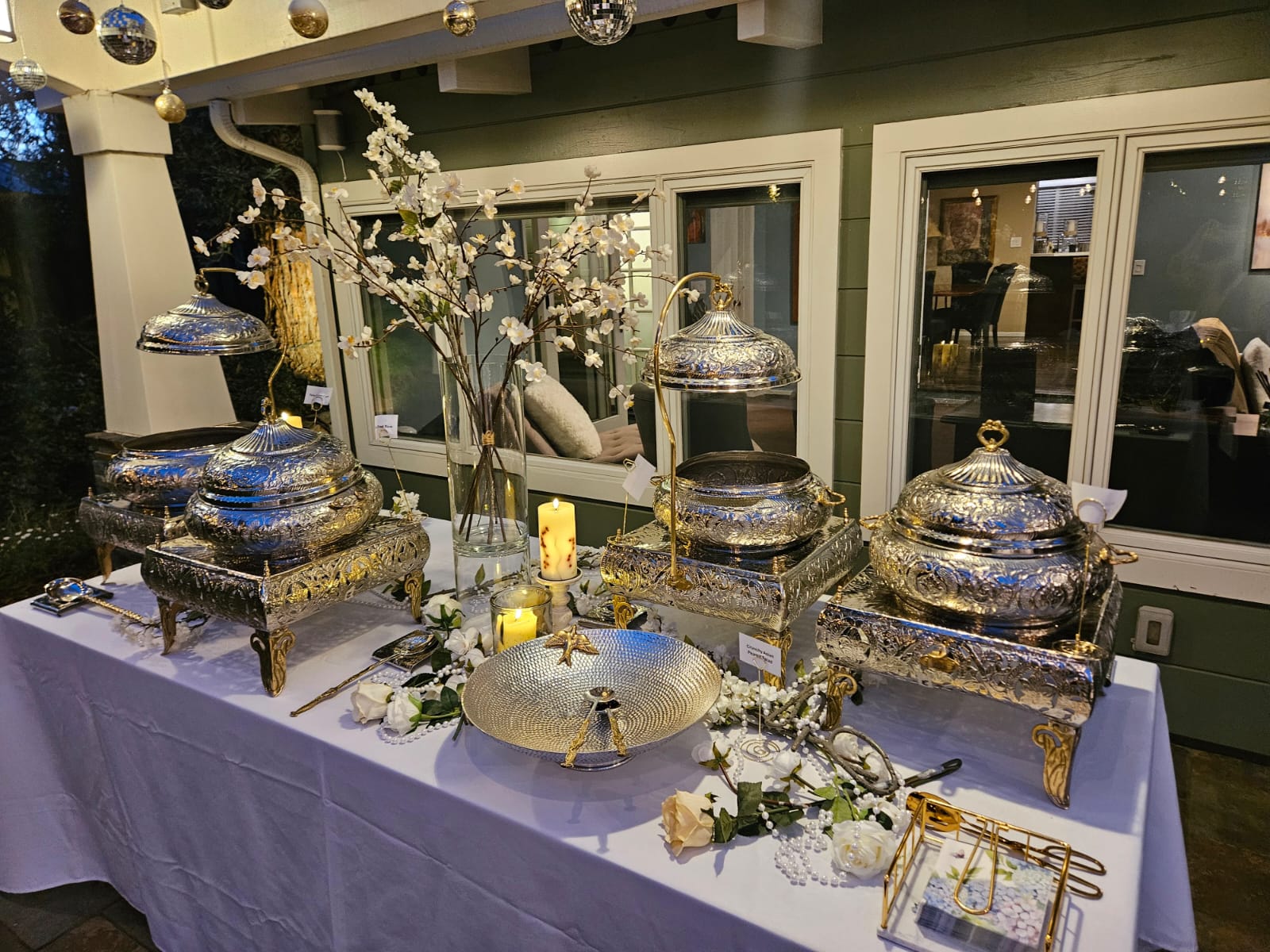 A candlelit buffet catered by Komal — engraved silver chafing dishes, blossoms, and string lights at dusk